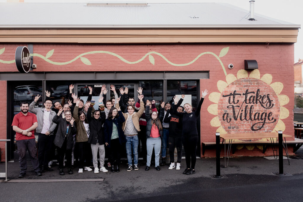A group of Hamlet's participants standing outside the cafe next to the wall mural that reads 'It takes a Village'. The participants are smiling for a photo with their hands in the air.