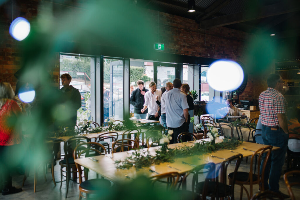 A group of people smiling and talking are walking into the Hamlet cafe. The cafe is set up for an event with bunches of white flowers and green foliage on the tables.