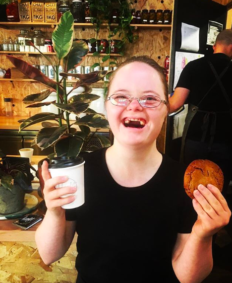 Hamlet's participant Alex is smiling for a photo at the counter wearing a black t-shirt and glasses and holding onto a takeaway cup and cookie.