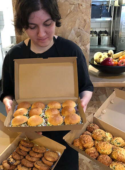 Hamlet's participant Amy is wearing a black t-shirt and apron, looking down at multiple cardboard trays filled with slider burgers, savoury muffins and cookies.