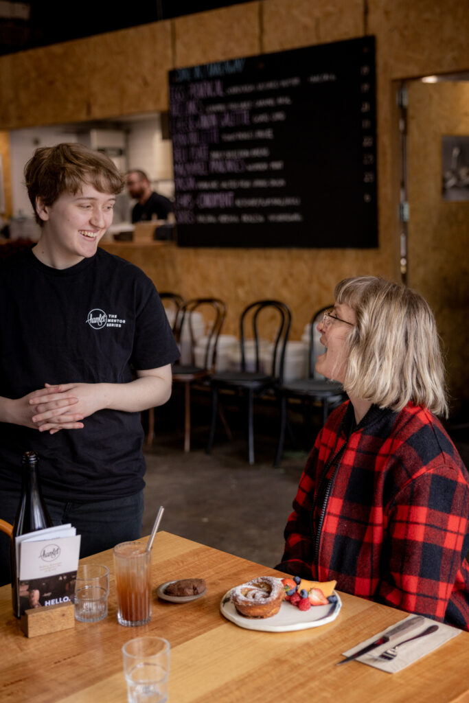 A Hamlet participant talking to a customer that's sitting down at a table in the cafe with a plate of sweets.