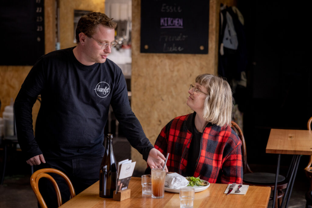 A Hamlet participant wearing a black t-shirt is talking to a customer seated in the cafe. The customer has just been served a burger and salad, and a large glass of cola.