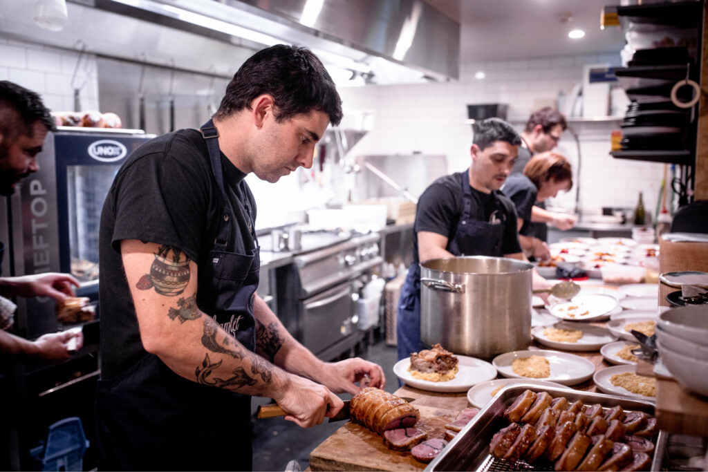 Five of Hamlet's chefs working in the kitchen wearing black t-shirts and navy aprons. One of the chefs is cutting up a rolled pork whilst another three chefs are working together plating up the meals.