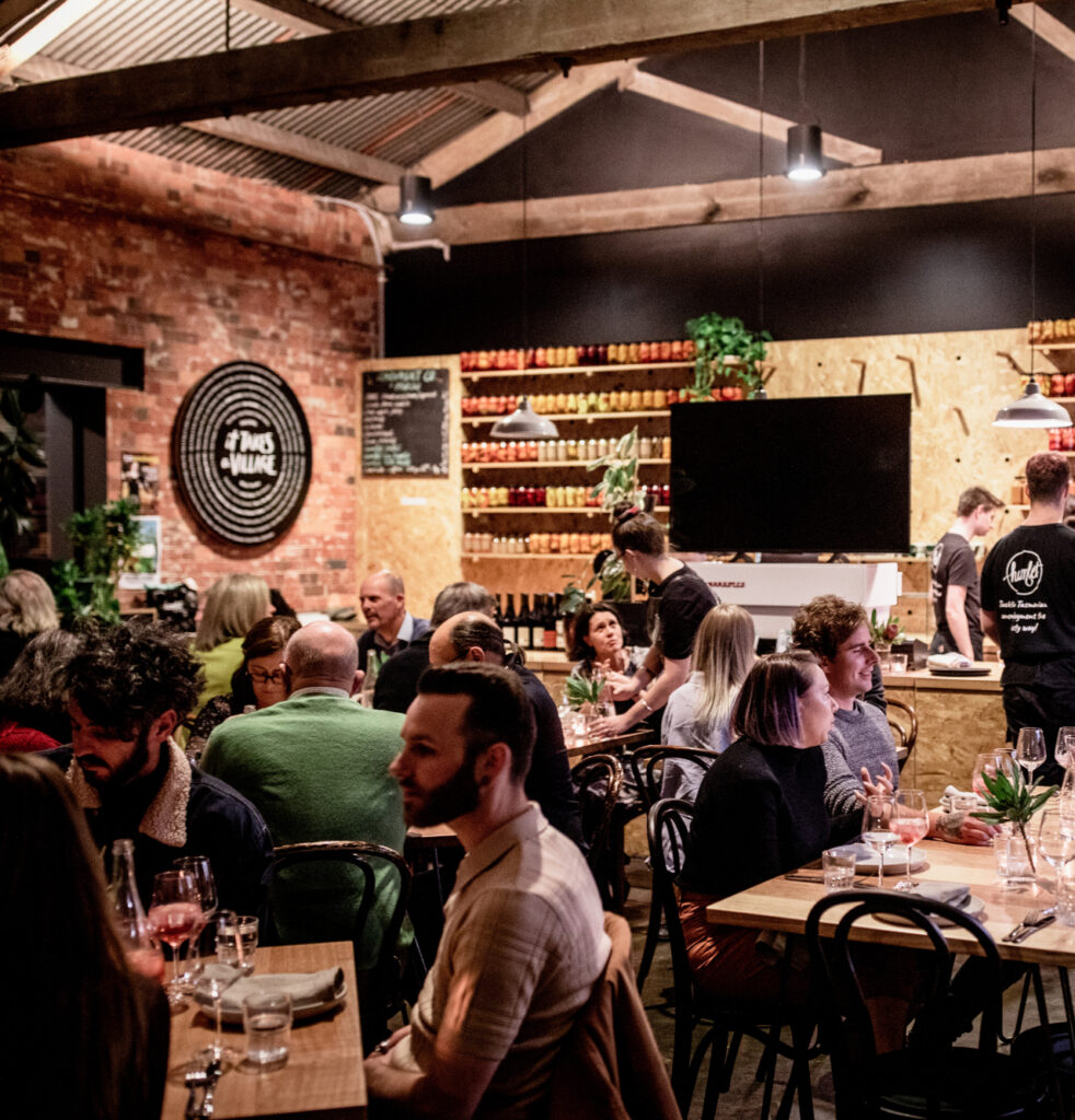 A large group of people seated at multiple tables in the Hamlet cafe. The people are talking and smiling. There are three Hamlet participants wearing black t-shirts working in the background.