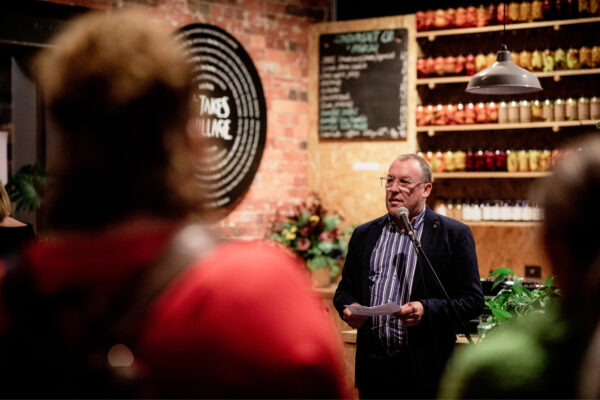 Person wearing a shirt and jacket standing in front of a microphone in the Hamlet cafe talking to a group of people.