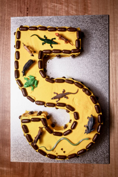 Number 5 birthday cake with yellow icing, chocolate liquorice bullets and small plastic animal figurines sitting on a foil plate.