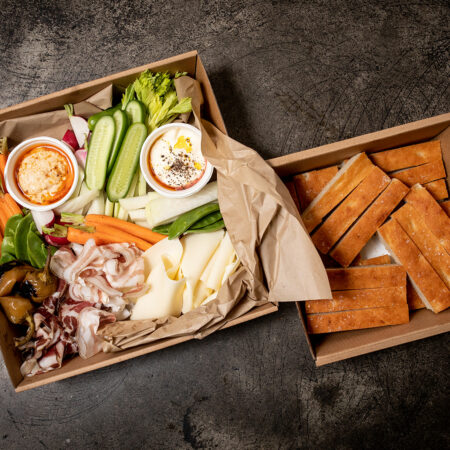 Large cardboard hamper filled with an array of deli meats, cucumber, carrots, snow peas and dips. There is a smaller cardboard hamper beside it filled with sliced bread.