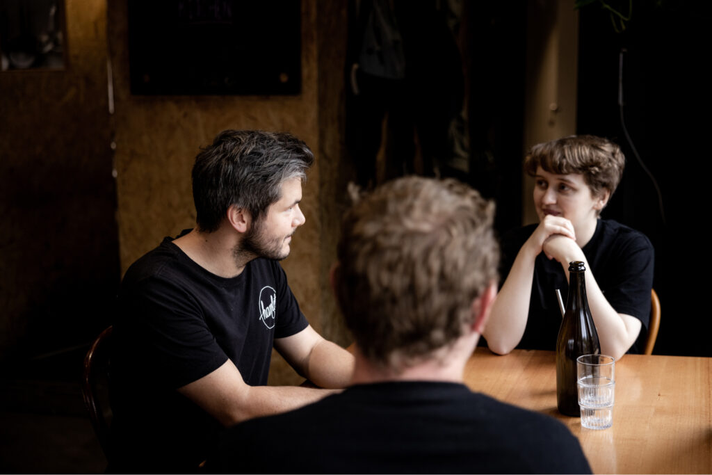 Three of Hamlet's participants are seated at a table in the cafe talking to each other wearing black t-shirts. There is a bottle of water and two glasses sitting on the timber table.