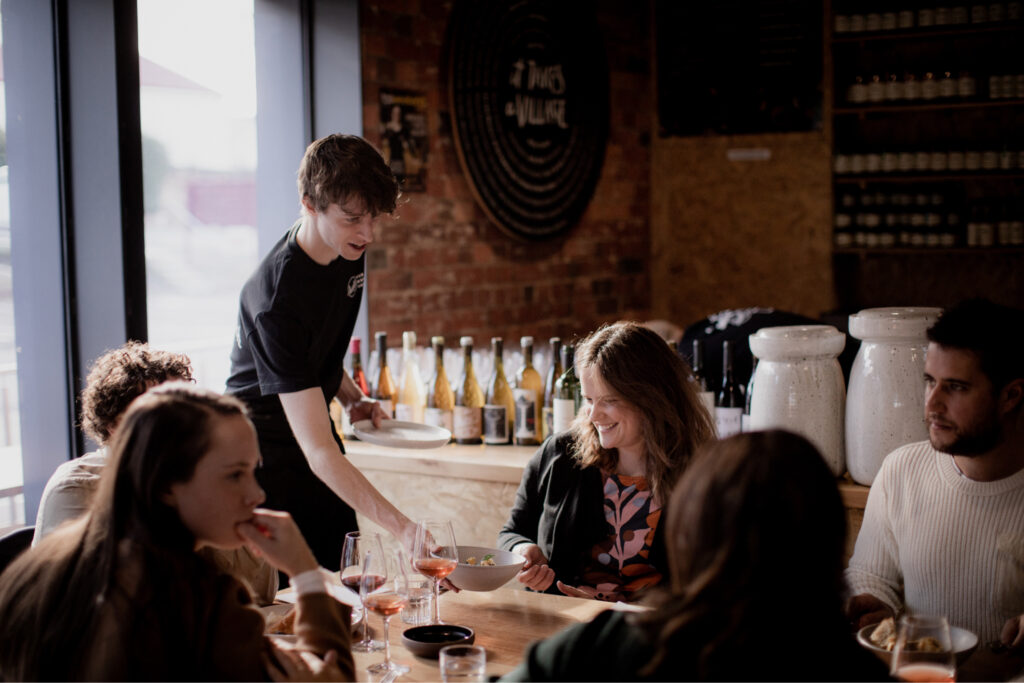 A Hamlet participant wearing a black t-shirt is serving a bowl of food to a customer seated at a table. There are another four customers seated at the same table talking to one another.