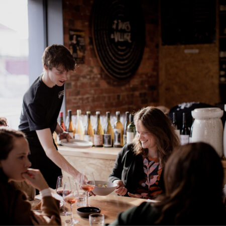 A Hamlet participant wearing a black t-shirt is serving a bowl of food to a customer seated at a table. There are another four customers seated at the same table talking to one another.