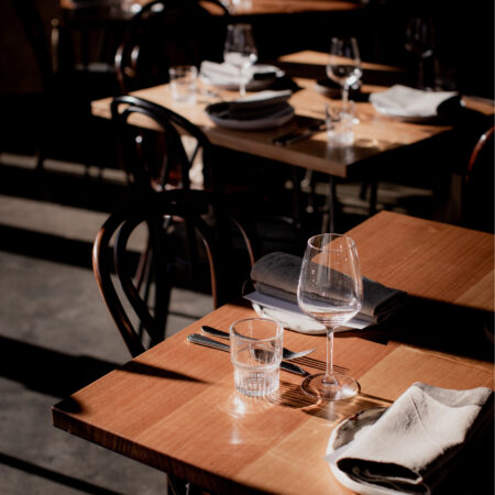 Three timber tables with chairs in the Hamlet cafe. There are a set of plates, napkins, cutlery, wine glasses and water glasses sitting on top of each table.