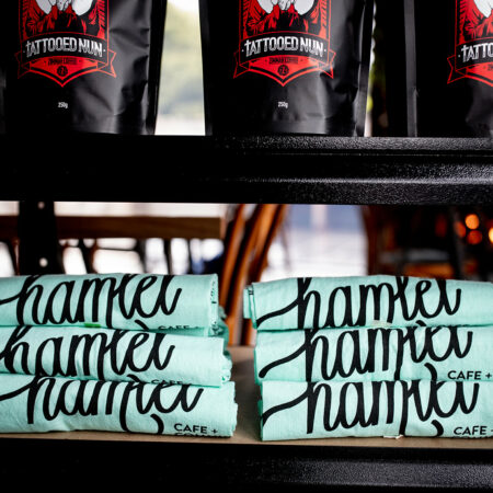Multiple pale green tea towels with black text that reads Hamlet Cafe and Community. The tea towels are stacked together on a black shelf in the cafe.