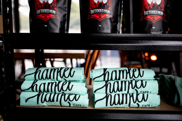 Multiple pale green tea towels with black text that reads Hamlet Cafe and Community. The tea towels are stacked together on a black shelf in the cafe.