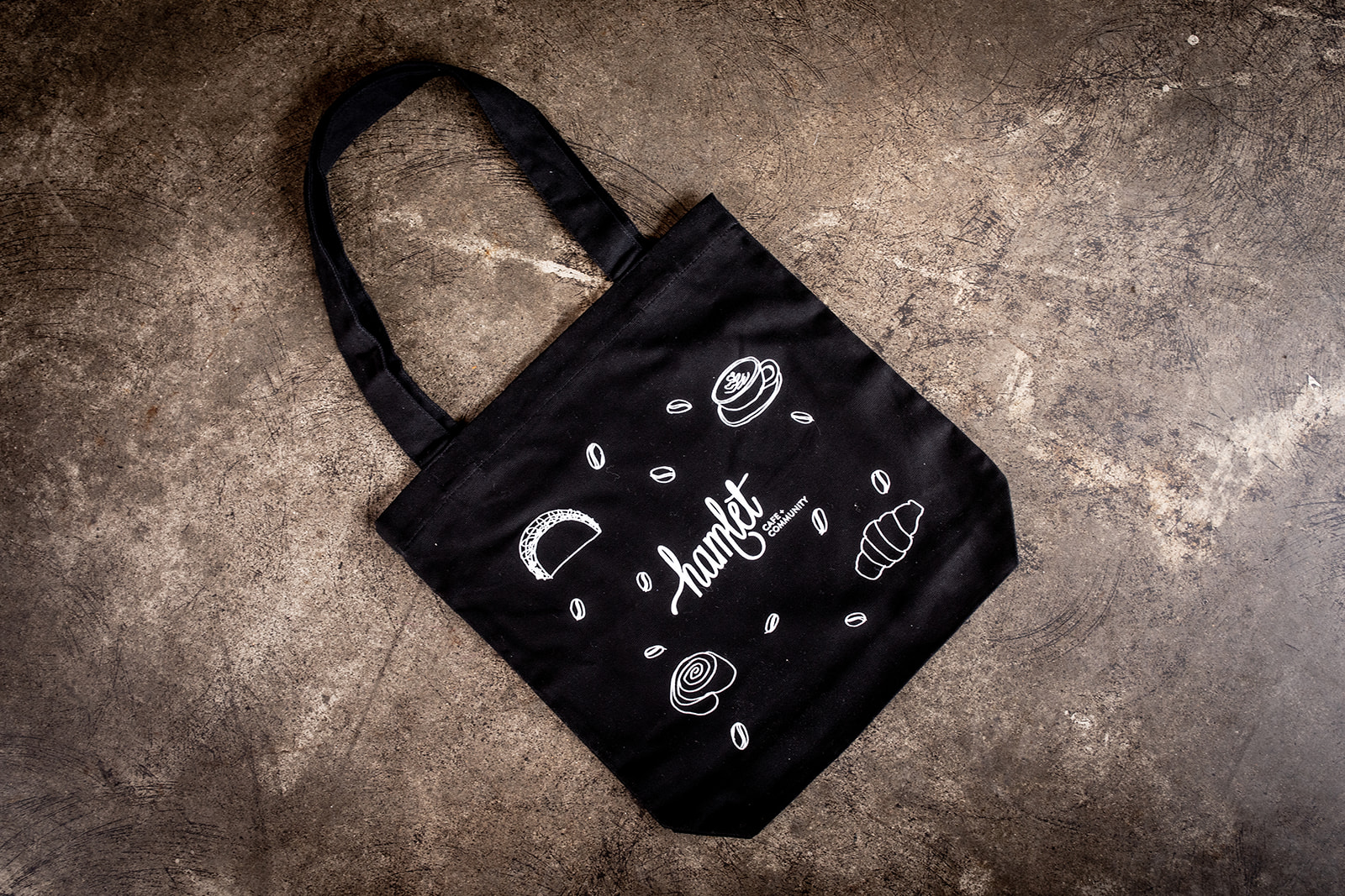 A black tote bag with white print, featuring the text Hamlet Cafe and Community surrounded by simple line drawings of cafe food.