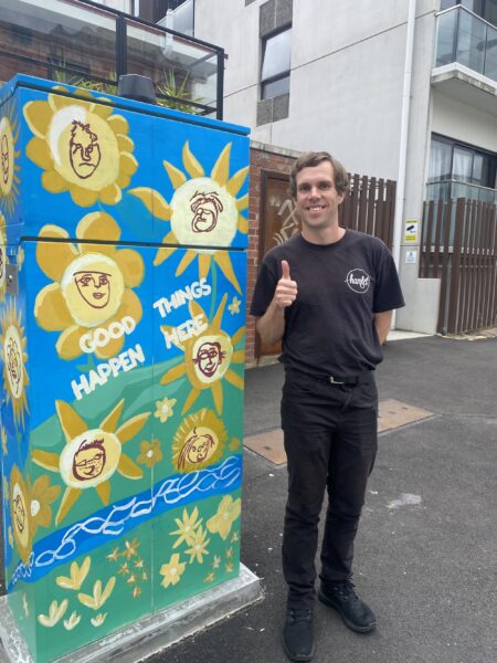 Hamlet's participant Luke standing beside a painted signal box with his thumbs up smiling for a photo wearing a black Hamlet t-shirt.