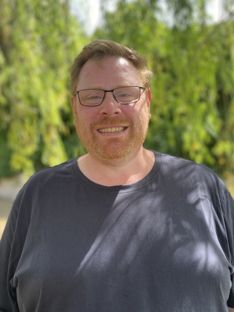 Hamlet's participant Charles standing in front of green foliage smiling for a photo wearing a black t-shirt.