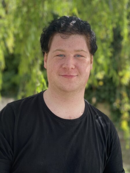 Hamlet's participant Liam standing in front green foliage, smiling for a photo wearing a black t-shirt.