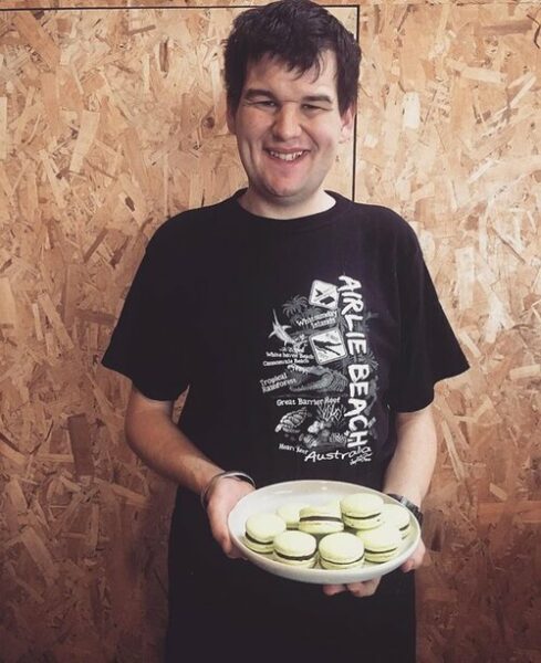 Hamlet's participant Adam is smiling for a photo standing in front of a chipboard wall, wearing a black t-shirt and holding a plate of macarons.