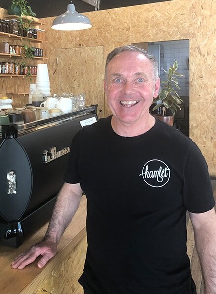 Hamlet's participant Andy smiling for a photo in front of the coffee machine wearing a Hamlet black t-shirt.