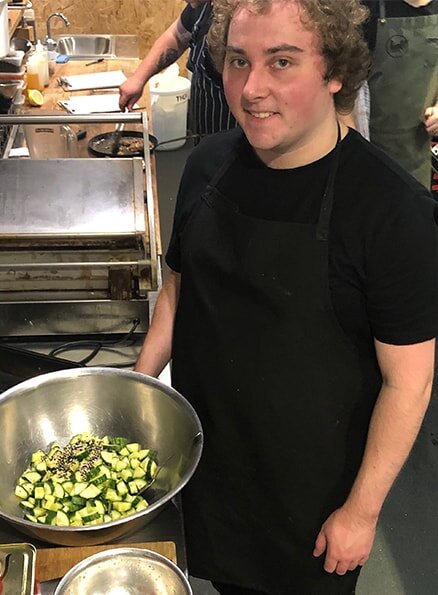 Hamlet's participant Liam is smiling for a photo in the kitchen standing next to a large bowl of chopped cucumber.