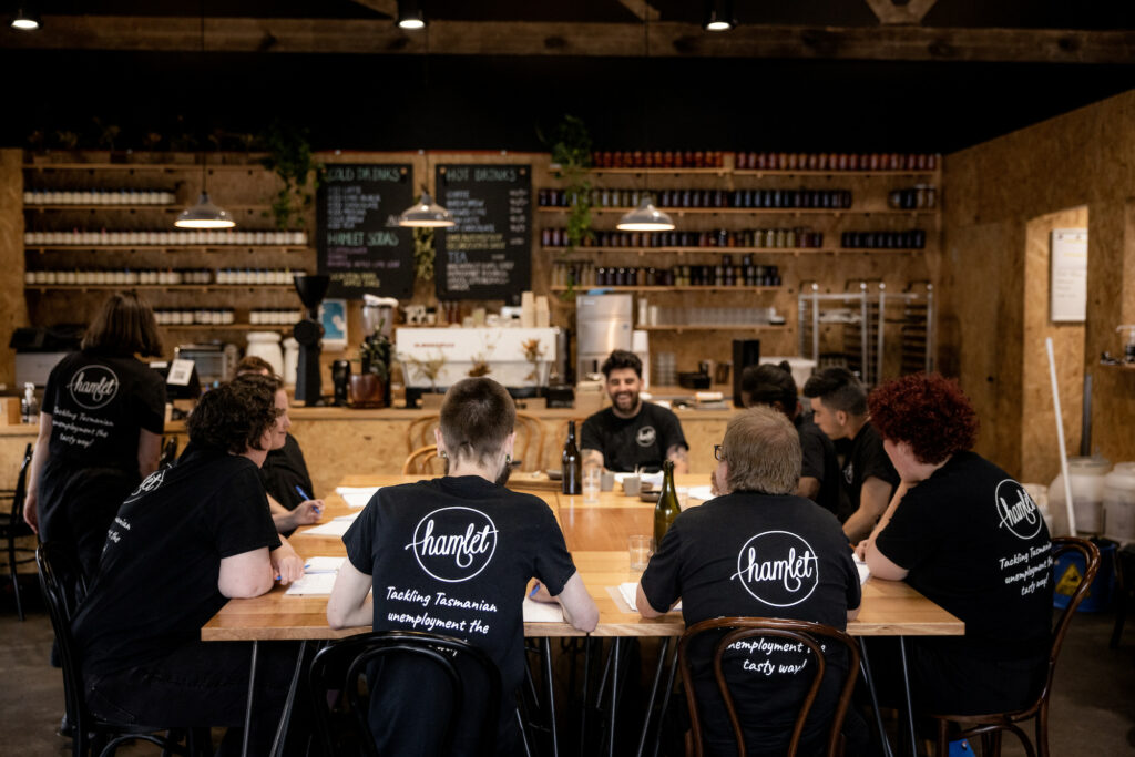 Group of Hamlet's participants sitting around a large table in the cafe taking notes on paper and wearing black Hamlet t-shirts.