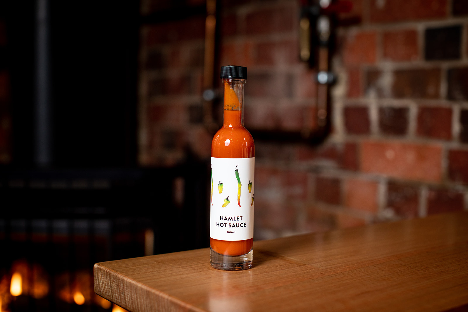 A bottle of Hamlet's house made Hot Sauce sitting on a timber table against a brick wall.