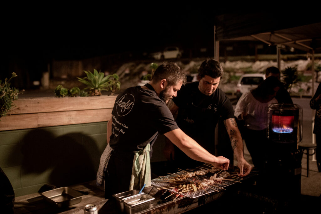 Two Hamlet chefs cooking skewers over an open grill outside wearing black t-shirts.