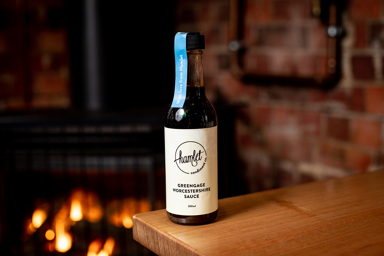 A bottle of Hamlet's house made Greengage Worcestershire Sauce sitting on a timber table against a brick wall.