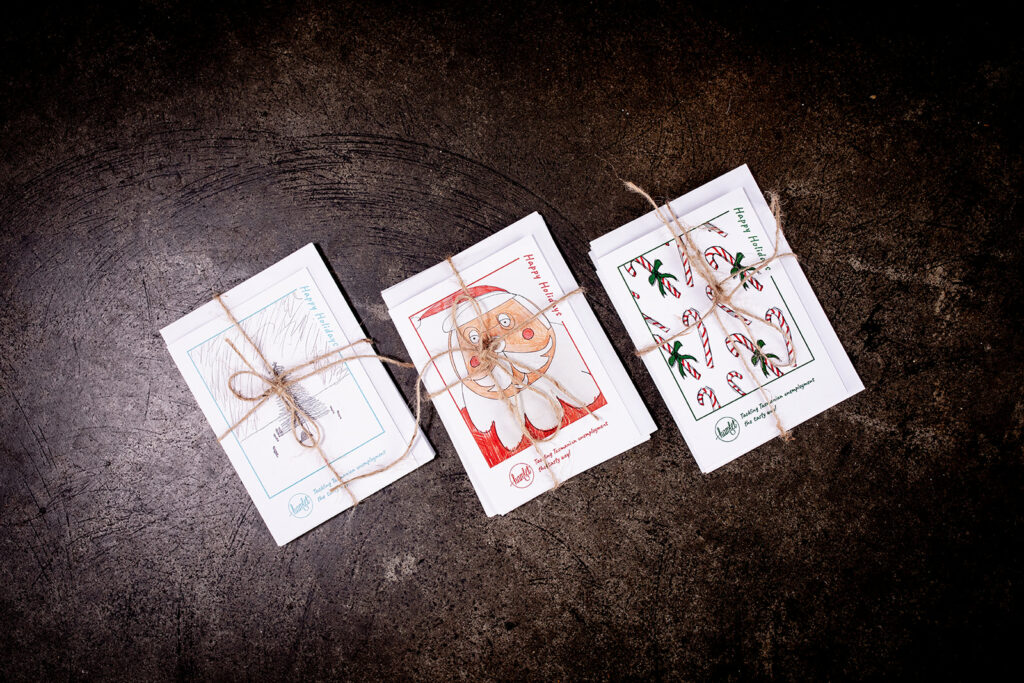 Three Hamlet holiday gift cards sitting on a concrete bench. Each card has artwork by Hamlet work experience participants and tied together in a bow with brown twine.