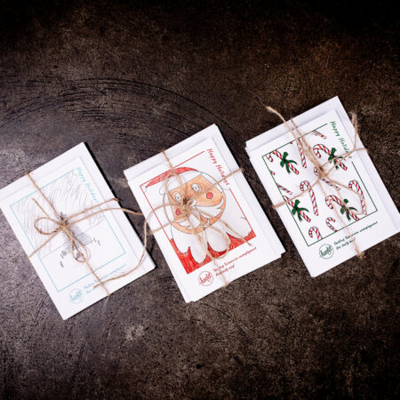 Three Hamlet holiday gift cards sitting on a concrete bench. Each card has artwork by Hamlet work experience participants and tied together in a bow with brown twine.