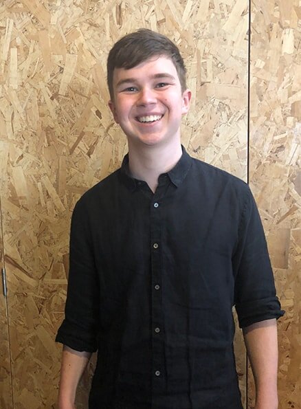 Hamlet's participant Jack smiling for a photo in front of a chipboard wall wearing a black shirt.