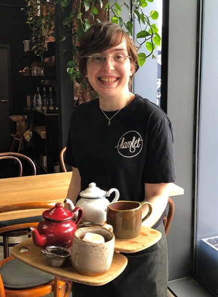 Hamlet's participant Keiran smiling for a photo holding two wooden serving boards with a tea pot and mug sitting on top of each.