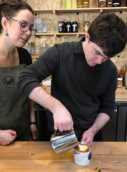 Hamlet's participant Liam standing at the counter pouring a jug of frothed milk into a cup of coffee. A colleague wearing a Hamlet apron is standing beside Liam watching him make a cappuccino.